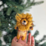 A handmade fluffy lion felt finger puppet is gently held by a female hand against a blurred Christmas tree background. This charming, handcrafted felt kids toy, made from soft, eco-friendly wool felt, showcases a golden mane, black eyes, black nose, and whiskers on its tan and brown body. This unique finger puppet is a fair-trade item, preserving traditional craftsmanship.