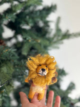 A handmade felt lion finger puppet is presented by a female hand against a blurred green Christmas tree backdrop. This unique felted finger puppet features a fluffy golden-yellow mane, a cream-to-yellow body with black paws, and a round face with stitched details. This cheerful felt toy for kids is handcrafted by Fair Trade artisans, promoting ethical and eco-friendly practices.