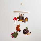 A Forest Friends wool felt baby mobile hangs against a plain white background, creating a gentle atmosphere. Handcrafted wool felt elements including an owl, fox, mushroom, hedgehog, acorn, and pine tree are suspended from a circular felt hoop by thin white strings. A red mushroom with white spots is to the left of the owl, with the fox hanging below them, and the hedgehog on the right. The mobile features soft browns, oranges, greens, and reds, evoking a warm woodland theme for a nursery.