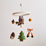A Forest Friends felt baby mobile, featuring handcrafted wool felt figures, is displayed against a plain white background. A gray owl, smiling orange fox, green pine tree, brown hedgehog, brown acorn, and charming mushroom are suspended by thin strings from a circular felt hoop, creating a calm, whimsical nursery scene.