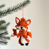 A handcrafted wool felt fox holding baby ornament is displayed on a pine branch against a bright white background. The orange mother fox, with a white chest and ear insides, lovingly embraces her smaller baby fox. Both felt foxes feature stitched closed eyes and petite black noses, with a red and white jute thread attached for hanging this charming Christmas ornament.