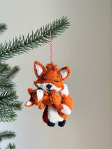 A heartwarming needle felted fox holding baby ornament hangs from a lush green Christmas tree branch, set against a neutral background. Crafted from soft orange and white wool felt, this unique felt ornament depicts a larger fox gently embracing a smaller one. Both foxes feature tiny perky ears, closed eyes, and subtle black stitched details, with a red and white jute thread attached for hanging.