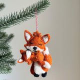 A heartwarming needle felted fox holding baby ornament hangs from a lush green Christmas tree branch, set against a neutral background. Crafted from soft orange and white wool felt, this unique felt ornament depicts a larger fox gently embracing a smaller one. Both foxes feature tiny perky ears, closed eyes, and subtle black stitched details, with a red and white jute thread attached for hanging.