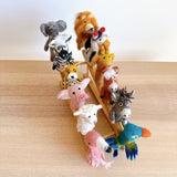 A playful set of 12 handmade wool felt finger puppets is displayed on a small bamboo stand, resting on a light wood surface against a white wall. This unique felt finger puppet collection features a gray elephant with large ears, a black and white zebra, a fluffy orange lion, and a black and white cow. Additional colorful characters include a yellow giraffe, spotted orange cheetah, pink pig, white wooly sheep, reddish-brown horse, gray donkey, pink flamingo, and blue and green parrot felt finger puppets.