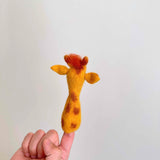 A needle felted giraffe finger puppet is playfully held on a female hand against a plain white background. This handcrafted felt kids toy features a long yellow wool felt neck with brown spots and an orange and yellow wool felt head detailed with ears. Its unique design brings a touch of wild fun to playtime.