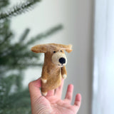 A handmade wool felt Golden Retriever finger puppet is held by a female hand, displayed against a blurred Christmas tree backdrop. The soft, golden-brown puppet features a black nose and a small stitched smile. Its handcrafted, needle-felted details create a lifelike, expressive look.