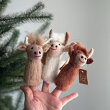 A cheerful wool felt Highland Cow finger puppet is displayed on a female hand, with a festive Christmas tree subtly blurred in the background. This charming, handmade felt finger puppet comes in light brown, white, and brown hues, each featuring distinct horns and a smiling face, perfect for engaging storytelling.