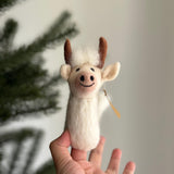 A handmade needle felted Highland Cow felt finger puppet is gently held by a female hand near a festive Christmas tree. Crafted from soft wool felt, this unique felt toy features a charming smiling face with a pink nose, black stitched details, fluffy white wool, and brown horns. A small tag is attached to its side, completing this delightful, handcrafted piece.