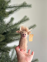 A female hand presents a charming needle felted Highland Cow finger puppet, set against a bright, airy background with a subtle Christmas tree branch. This handcrafted felt kids toy features a light brown body with a fluffy light brown fringe and curved white horns. A small tag is visible on the side of this unique finger puppet.