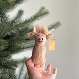 A female hand presents a charming needle felted Highland Cow finger puppet, set against a bright, airy background with a subtle Christmas tree branch. This handcrafted felt kids toy features a light brown body with a fluffy light brown fringe and curved white horns. A small tag is visible on the side of this unique finger puppet.