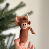 A playful, needle felted Highland cow finger puppet is held by a female hand, presented against a blurred Christmas tree background. This charming, handmade felt toy features brown fluffy hair, small ears, and curved horns, with black dot eyes and a stitched smile. A small tag is attached to its tail.