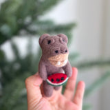 A handcrafted felt hippopotamus finger puppet, holding a watermelon slice, is delicately presented by a female hand in a bright, airy setting. This muted brown hippopotamus features small, rounded ears and tiny black eyes. The vibrant red watermelon slice it holds is detailed with black seeds and a thin green rind. Blurred greenery in the background suggests a playful, summery atmosphere.