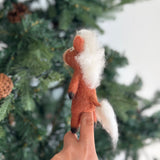 A handcrafted fair-trade needle felted horse finger puppet, made from 100% wool in Nepal, is held upright on a finger, presented against a blurred Christmas tree adorned with pine cones, creating a festive scene. This unique felt horse puppet features a tan body, a fluffy white mane and tail, and black hooves, perfect for imaginative storytelling.