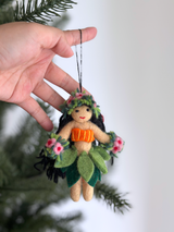 A Hula Girl Ornament is held by a female hand, prominently displayed against a lush green Christmas tree. This festive Christmas ornament features a light tan body, an orange top, and a dark green grass skirt. Adorned with a flower crown and flower bracelets, this charming ornament is ready to hang, bringing joyful, playful vibes to holiday decor.