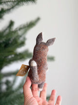 A brown wool felt kangaroo finger puppet is held in a hand, set against a soft, bright background with a blurred Christmas pine tree. This handcrafted puppet features pointed ears, a small tail, and lighter accents on its paws and ear insides, highlighting its charming, tactile design and eco-friendly craftsmanship. A small tag hangs from it, emphasizing its handmade origin.