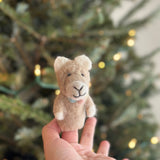 A handcrafted wool felt llama finger puppet is gently held by a female hand, set against a softly blurred Christmas tree background with twinkling lights. This light brown llama puppet features white ears, a cute smile, a soft, fuzzy texture, and a pastel pompom collar, creating a cozy and festive holiday scene.
