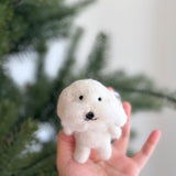 A needle felted Maltese dog finger puppet is held by a female hand against a blurred Christmas tree background. This charming, handmade felt toy features fluffy white hair, a black nose, and black eyes, perfect for imaginative play.