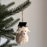 A handmade wool felt graduate ram ornament hangs gracefully on a green pine tree branch against a light grey background. This charming Christmas ornament features a fluffy cream coat, curled brown horns, and a miniature black graduation cap, with a natural twine string attached for easy display.