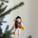 This charming handmade felt mouse family ornament dangles from a Christmas tree branch against a soft background. The mouse wears a black hat and a brown vest with buttons, clutching a bundle of wheat. A jute string secures this unique Christmas ornament, creating a cozy, festive display.