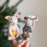 Two playful, handmade felt mouse finger puppets are held in a female hand, set against a soft-focus Christmas tree background. These approximately 4-inch tall, needle-felted creations are crafted in Nepal from 100% organic wool felt with eco-friendly dyes. One mouse is white and pink, holding a yellow cheese wedge, while the other is brown and gray with a white head covering. Both feature tiny whiskers and ears, adding to their charming, unique design.