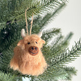 A Needle Felted Highland Cow ornament hangs gracefully on a green Christmas tree branch. This handmade wool felt ornament features a soft, light-brown texture, small white horns, and a sweet, smiling face with dark brown needle-felted details for the nose and eyes.