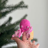 A handmade magenta pink wool felt octopus finger puppet is presented by a female hand against a pine tree backdrop. This unique, handcrafted felt puppet features eight lighter pink curled tentacles, poppy smiley eyes, and a charming smile. A small tag highlights its fair-trade origin, supporting female artisans.