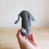 A needle felted old gray dog finger puppet is gently held by a female hand against a light, neutral backdrop, showcasing its artisanal craftsmanship. This handcrafted, 100% wool felt finger puppet appears predominantly gray with a lighter tail, viewed from its backside. Its tactile design highlights a playful and engaging appeal.
