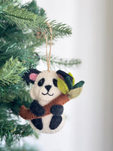 A charming wool felt panda ornament hangs against a festive Christmas tree backdrop. This delightful panda is white with black ears and body accents, featuring a pink inner ear. It playfully clutches a green-leafed bamboo stalk and is suspended by a jute string, ready to add a touch of handmade charm.