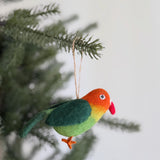 A vibrant wool felt parrot ornament hangs cheerfully on a green Christmas tree branch. This unique Christmas ornament features the parrot's head transitioning from orange to yellow, with a green back, tail, and orange feet. A red beak and round eye complete its design, showcasing the visible wool felt texture of this handcrafted piece.