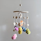 A Pastel Sheep Wool Felt Baby Mobile hangs from a white ceiling, featuring a cat figurine, moon, and multiple pastel-colored sheep in pink, blue, green, yellow, and purple. The mobile is handcrafted from soft wool felt and suspended from a felt hoop for gentle movement.