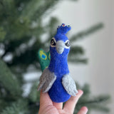 A handcrafted needle-felted peacock finger puppet is held gently in a female hand, prominently displayed against a blurred Christmas tree and soft white wall background. This unique wool felt finger puppet features a predominantly blue body with a grey beak, grey wings, and a distinctive green and blue tail fan, showcasing its ethical craftsmanship.