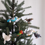 A set of handmade felt bird ornaments hangs on a festive Christmas tree, set against a simple white background. The bird ornament set includes a parrot, a pigeon, a dove with olive twig, a seagull, a blue budgerigar, and a white-throat sparrow. These unique, handcrafted felt ornaments are made from natural wool felt, featuring soft tones and lifelike needle-felted details with visible feather patterns. Each one is suspended by a rustic jute string, which adds a whimsical touch to holiday decor.