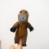A needle felted wool platypus finger puppet rests on a female finger against a plain white background. The handcrafted felt toy features a brown body, a grey bill, and black feet, highlighting its detailed 3D design.