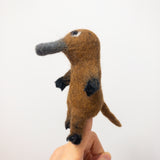 A handcrafted wool felt platypus finger puppet is displayed on a person's finger against a clean white background. This unique needle felted puppet is primarily brown with black accents on its feet and hands. It features a distinctive gray bill and a tail, showcasing its charming 3D-designed shape, ideal as a kids' toy.