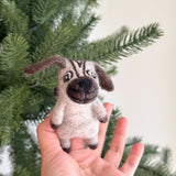 A handmade felt pug finger puppet is held by a female hand, positioned in front of a soft-focus Christmas pine tree. This charming felt pug finger puppet is light beige, featuring characteristic brown wrinkled markings on its face. It also has floppy brown ears, a round dark brown nose, and black eyes, intricately capturing the beloved pug's distinct features.