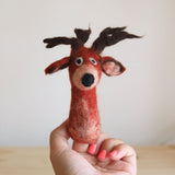 A handmade needle-felted red reindeer finger puppet is held upright in a female hand against a light background. The intricately crafted reindeer features a rounded red nose, small white and black eyes, and brown felt antlers. Its body is primarily red with lighter, mottled accents, showcasing the textured needle-felted material. This unique felt toy for kids is presented in a close-up studio shot, highlighting its whimsical design.