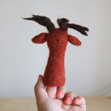 A red reindeer wool felt finger puppet is held upright by a female hand against a plain, light background. This charming handmade felt toy for kids features a vibrant red body, a cute red nose, and darker brown antlers. The hand, with coral nail polish, showcases the puppet's soft texture and unique craftsmanship.