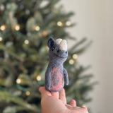 A handmade needle-felted rhino finger puppet is gently held in a woman's hand, set against a softly lit Christmas tree backdrop. This unique rhino puppet features a gray body with a pale pink chest and a white horn. Its intricate, handcrafted details and soft, fuzzy texture make it perfect for imaginative play and storytelling.