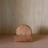 Handcrafted Bali rattan coaster set of 5, handmade in Bali, is presented neatly inside its matching rectangular woven holder. The warm, golden-brown rattan set is centered on a light wood shelf, creating a beautiful tonal harmony with the pale, vertical wood-grain background. The soft lighting highlights the intricate sunburst weave of the front Bali rattan coaster, evoking a feeling of serene, minimalist elegance and showcasing the product's natural, artisanal quality.