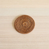 Handcrafted Bali rattan coaster, one from a set of 5 handmade in Bali, is beautifully displayed in a minimalist top-down shot. The single, golden-brown coaster is centered on a light, fine-grained wood surface, its intricate sunburst weave creating a striking textural focal point. The clean, simple environment enhances the coaster's natural, artisanal feel, evoking a sense of calm and organic elegance.