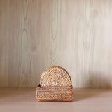 Handcrafted Bali rattan coaster set of 5, handmade in Bali, is presented neatly inside its matching rectangular woven holder. The warm, golden-brown rattan set is centered on a light wood shelf, creating a beautiful tonal harmony with the pale, vertical wood-grain background. The soft lighting highlights the intricate sunburst weave of the front Bali rattan coaster, evoking a feeling of serene, minimalist elegance and showcasing the product's natural, artisanal quality.