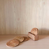 Handcrafted Bali rattan coaster set of 5, handmade in Bali, is artfully arranged on a light wood shelf against a warm, wood-grain background. Three golden-brown coasters are fanned out in the foreground, showcasing their intricate sunburst weave, while the other two sit in their rectangular woven holder to the right. The scene evokes a feeling of natural, rustic elegance, highlighting the beauty of the Bali rattan coaster set in a serene, minimalist environment.