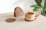Handcrafted Bali rattan coaster set of 5, handmade in Bali, is displayed in a bright and serene setting on a light wood table. One dark-toned Bali rattan coaster is in use, holding a beautifully carved wooden mug, while another lies flat nearby. In the background, the matching holder contains the rest of the set, with a soft-focus green plant adding a touch of nature. The scene evokes a peaceful, organic feeling, showcasing the coaster's practical function and rustic elegance in a calm, modern home.