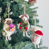 The Santa's Little Helper Sheep Ornament, a handcrafted wool felt Christmas ornament, hangs suspended by a jute string from a Christmas tree branch. This snowy white sheep ornament cradles a miniature Santa doll, showcasing its soft, textured appearance and adding rustic charm to the festive holiday decor. A simple white wall provides a clean backdrop.