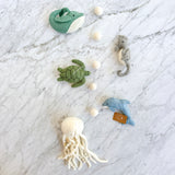 A colorful felt sea life baby mobile, presented as a garland, rests on a white marble surface. It features five detailed felt sea creatures: a green and white whale, a green sea turtle, a gray seahorse, a light blue dolphin, and a white jellyfish. White felt balls connect the figures along the string, adding a playful touch.