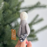 A handmade felt seagull finger puppet, worn on a female hand, is set against a blurred Christmas pine tree background. This unique seagull puppet features a white head, a light gray body, and a darker gray tail with white dots, embodying fine craftsmanship.