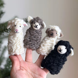 A handmade felt sheep finger puppet is presented on a female hand against a blurred backdrop of green foliage, evoking a playful and cozy atmosphere. This unique felted finger puppet features fluffy, textured wool, small ears, and a simple embroidered face, adding to its whimsical charm and inviting creative play.