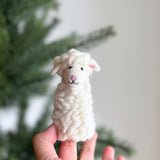 A fluffy white handmade wool felt sheep finger puppet is held by a hand against a blurred green Christmas tree backdrop. This charming sheep puppet features a textured body, small perky ears with pink interiors, tiny black eyes, and a pink stitched nose, creating a cozy and playful feel.