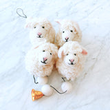 A charming felt sheep garland is displayed on a white marble surface, creating a cheerful presentation. It features three plush, off-white wool felt sheep, each with sweet pink accents on its nose and inner ear. A thin black string connects the sheep, interspersed with round, white felt balls, and ending with a shiny copper bell. The garland's fluffy texture evokes warmth and handmade charm.