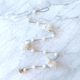 A charming wool felt sheep garland is displayed on a bright, marbled surface. It features three-dimensional white felt sheep figures connected by a thin black string with interspersed white felt balls. A whimsical copper bell dangles from one end.